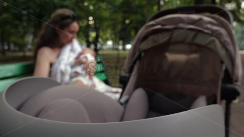 Close up of a stroller with a mother breastfeeding her baby while sitting on a park bench in the background - Starpik Stock