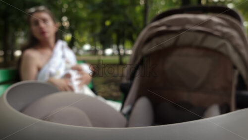 Close up of a stroller with a mother breastfeeding her baby while sitting on a park bench in the background - Starpik Stock