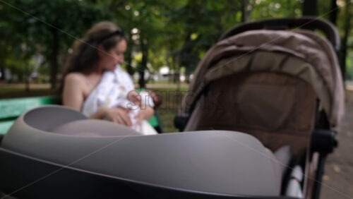 Close up of a stroller with a mother breastfeeding her baby while sitting on a park bench in the background - Starpik Stock