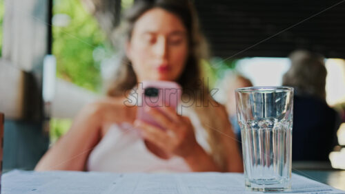 Close up of an empty glass on a table with a blurred view of a woman scrolling on her phone at a restaurant - Starpik Stock