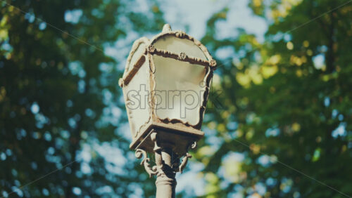 Close up of an ornate vintage street lamp against green trees on a sunny day - Starpik Stock