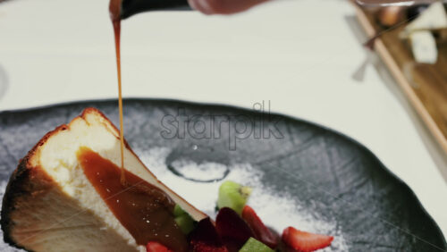 Close up of caramel sauce being poured over a slice of cheesecake served with fresh strawberries and kiwi, garnished with powdered sugar on a plate at a restaurant - Starpik Stock