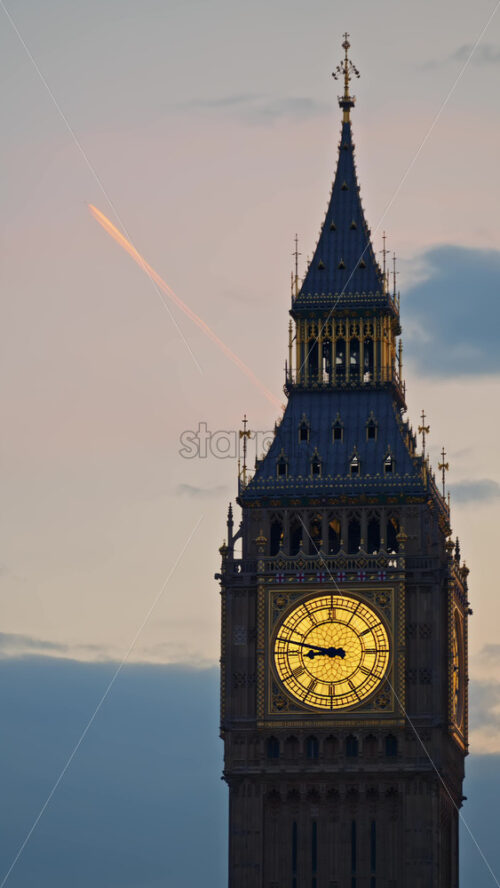 Close-up view of Big Ben illuminated at dusk with airplane passing by in London, England. Vertical - Starpik Stock