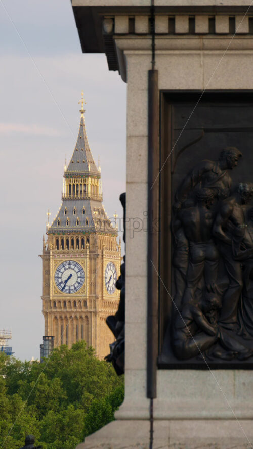 Close-up view of the Big Ben clock tower illuminated by golden sunlight, partly framed by a historic monument in London, England. Vertical - Starpik Stock