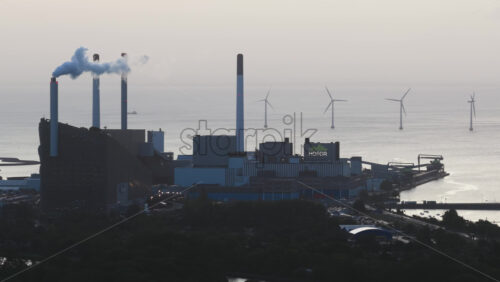 Copenhagen, Denmark – August 3, 2025: Aerial drone view of CopenHill energy plant with smoking chimneys, overlooking the sea and offshore wind farms. Early morning, dramatic light - Starpik Stock