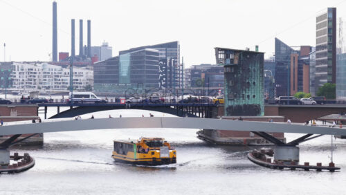 Copenhagen, Denmark – August 4, 2025: Aerial drone view of a yellow Copenhagen harbor bus passing under Lille Langebro pedestrian bridge, with cyclists crossing above - Starpik Stock