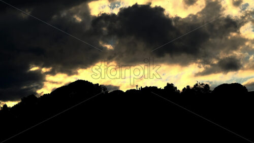 Dark clouds drift over a glowing yellow sky above a hilltop - Starpik Stock