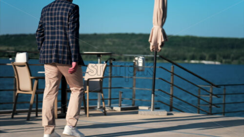 Elegantly dressed man walking on a scenic terrace with a view of a calm body of water - Starpik Stock