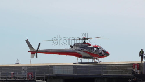 Fontvieille, Monaco – July 9, 2025: Red-silver helicopter idling on the Monaco Heliport with ground crew in hi-vis vests beside the pier - Starpik Stock