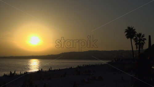 Golden sunset over the Mediterranean Sea with silhouettes of palm trees and people relaxing on the beach in Nice, France - Starpik Stock
