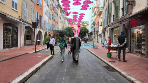 Grasse, France – August 10, 2025: Picturesque street decorated with pink umbrellas hanging above, surrounded by charming boutiques and people strolling - Starpik Stock