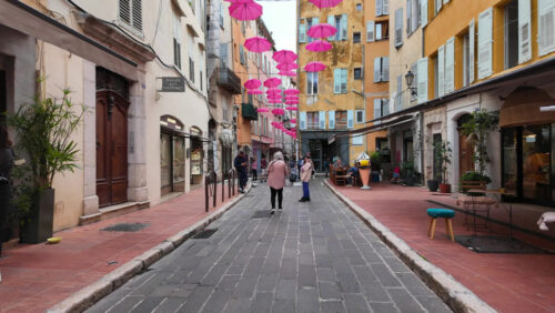 Grasse, France – August 10, 2025: Picturesque street decorated with pink umbrellas hanging above, surrounded by charming boutiques and people strolling - Starpik Stock