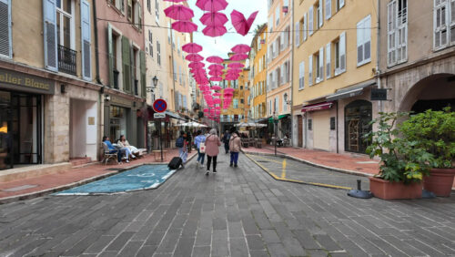 Grasse, France – August 10, 2025: Picturesque street decorated with pink umbrellas hanging above, surrounded by charming boutiques and people strolling - Starpik Stock