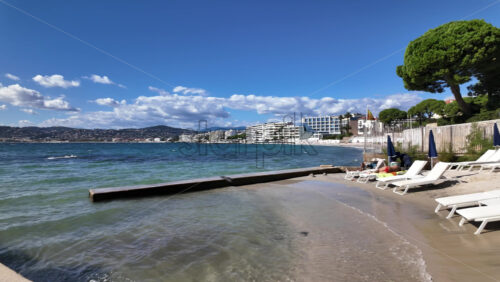 Juan-les-Pins, France – July 12, 2025: Sunbathers lounge under yellow umbrellas as gentle waves roll onto a sandy urban beach on a clear day - Starpik Stock