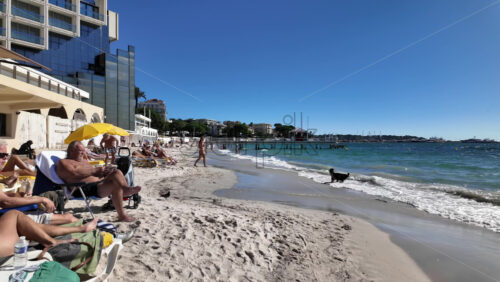 Juan-les-Pins, France – July 12, 2025: Sunbathers lounge under yellow umbrellas as gentle waves roll onto a sandy urban beach on a clear day - Starpik Stock