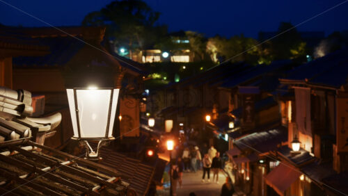 Kyoto, Japan – April 11, 2025: Close up of a street lamp with a blurred view of people walking on the Ninenzaka stone-paved pedestrian road, lined with traditional buildings and shops in the evening - Starpik Stock