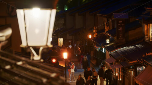 Kyoto, Japan – April 11, 2025: Close up of a street lamp with a view of people walking on the Ninenzaka stone-paved pedestrian road, lined with traditional buildings and shops in the evening - Starpik Stock