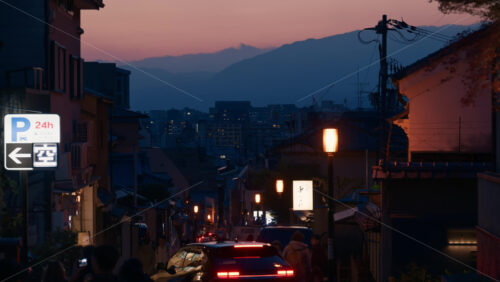 Kyoto, Japan – April 11, 2025: People and cars moving through Ninenzaka stone-paved road, lined with traditional buildings and shops at sunset, with mountains on the background - Starpik Stock