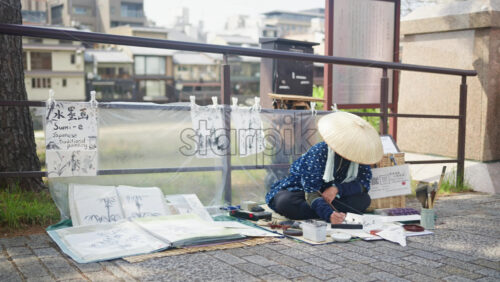 Kyoto, Japan – April 11, 2025: Person wearing a traditional Japanese hat painting Sumi-e traditional style in black and white on the street of Gion district - Starpik Stock