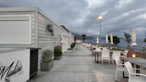 La Condamine, Monaco – July 18, 2025: Empty white chairs and closed umbrellas line a waterfront terrace as storm clouds roll in, cruise ship and city lights beyond - Starpik Stock