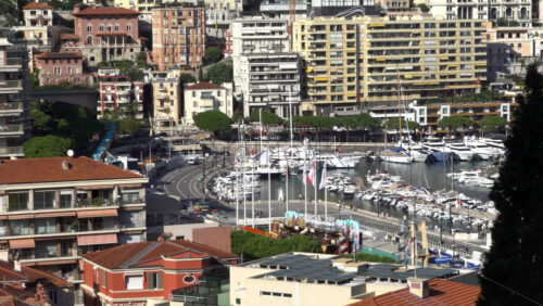 La Condamine, Monaco – July 4, 2025: Busy curve of Quai Albert-1er with traffic, Monaco flags, a seasonal funfair, and rows of yachts moored in Port Hercule below residential blocks - Starpik Stock
