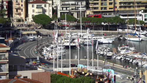 La Condamine, Monaco – July 4, 2025: Busy curve of Quai Albert-1er with traffic, Monaco flags, a seasonal funfair, and rows of yachts moored in Port Hercule below residential blocks - Starpik Stock