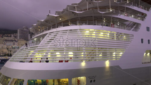 La Condamine, Monaco – July 4, 2025: Evening view of a cruise ship docked in Monaco, with illuminated decks and the city skyline in the background under a cloudy sky - Starpik Stock