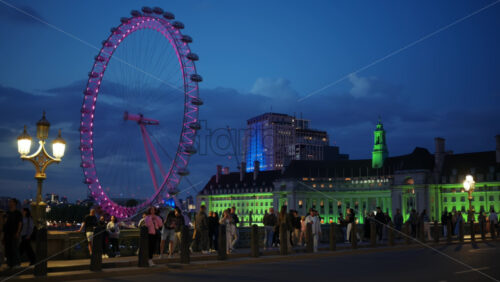 London, England – May 11, 2025: Crowds walking along Westminster Bridge with the London Eye glowing pink and County Hall lit in green in the evening - Starpik Stock