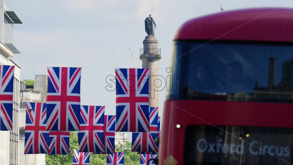 London, England – May 11, 2025: Red double-decker bus driving past Union Jack banners with the Palace of Westminster and Nelson’s Column in the background - Starpik Stock