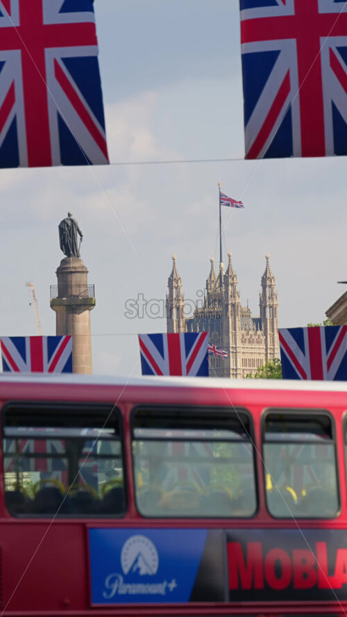 London, England – May 11, 2025: Red double-decker bus driving past Union Jack banners with the Palace of Westminster and Nelson’s Column in the background. Vertical - Starpik Stock