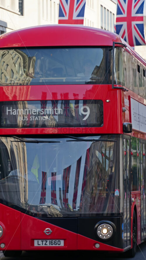 London, England – May 11, 2025: Red double-decker bus driving under moving under multiple flags of the United Kingdom hanging proudly above a busy city street in the city center. Vertical - Starpik Stock
