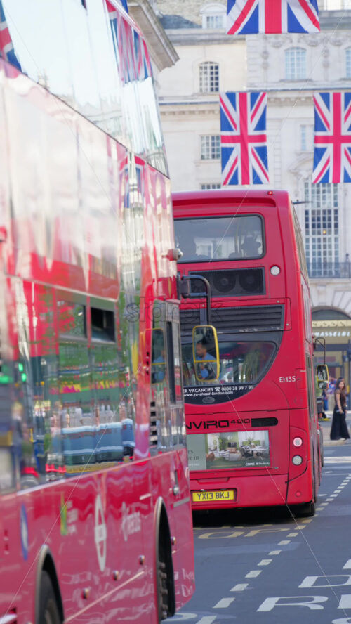 London, England – May 11, 2025: Red double-decker bus driving under moving under multiple flags of the United Kingdom hanging proudly above a busy city street in the city center. Vertical - Starpik Stock