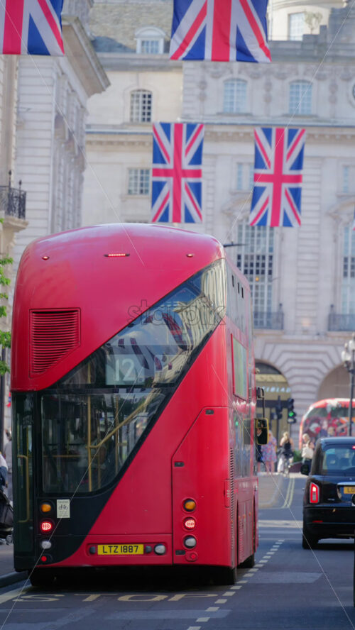 London, England – May 11, 2025: Red double-decker bus driving under moving under multiple flags of the United Kingdom hanging proudly above a busy city street in the city center. Vertical - Starpik Stock