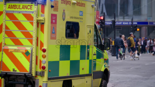 London, England – May 12, 2025: Ambulance with flashing lights driving through a busy intersection surrounded by pedestrians and vehicles - Starpik Stock
