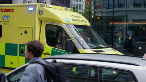 London, England – May 12, 2025: Ambulance with flashing lights driving through a busy intersection surrounded by pedestrians and vehicles - Starpik Stock