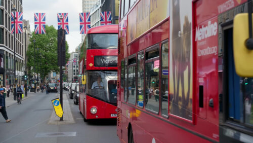 London, England – May 12, 2025: Classic red double-decker buses, cars and people moving on the street under Union Jack flags - Starpik Stock