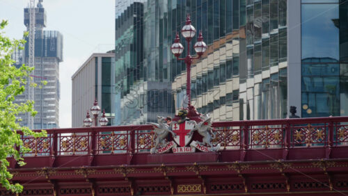 London, England – May 12, 2025: Double-decker bus moving on a red bridge with a dragon crest and with modern glass skyscrapers in the background - Starpik Stock