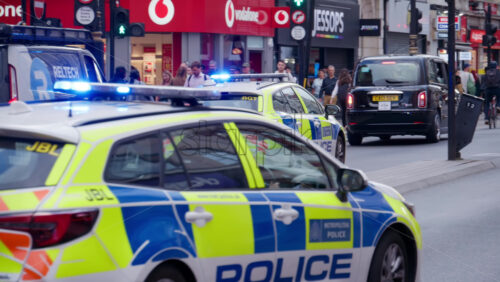 London, England – May 12, 2025: Two UK police cars with flashing blue lights drive through a busy street, passing pedestrians and cyclists during the day - Starpik Stock