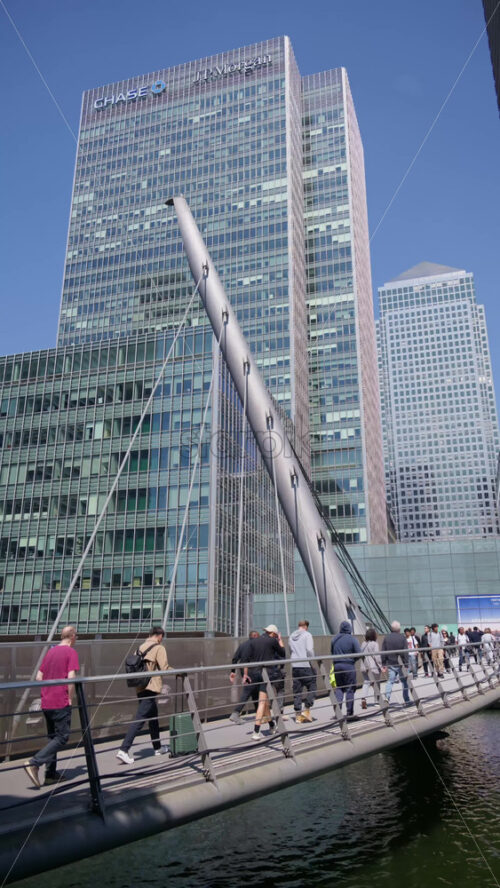 London, England – May 13, 2025: Canary Wharf skyscrapers with people walking across a sleek pedestrian bridge. Vertical - Starpik Stock