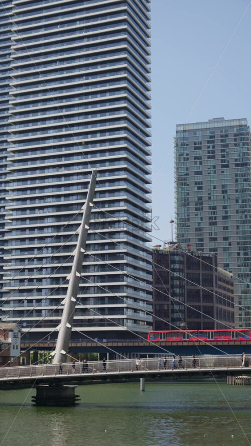 London, England – May 13, 2025: Canary Wharf skyscrapers with people walking across a sleek pedestrian bridge. Vertical - Starpik Stock