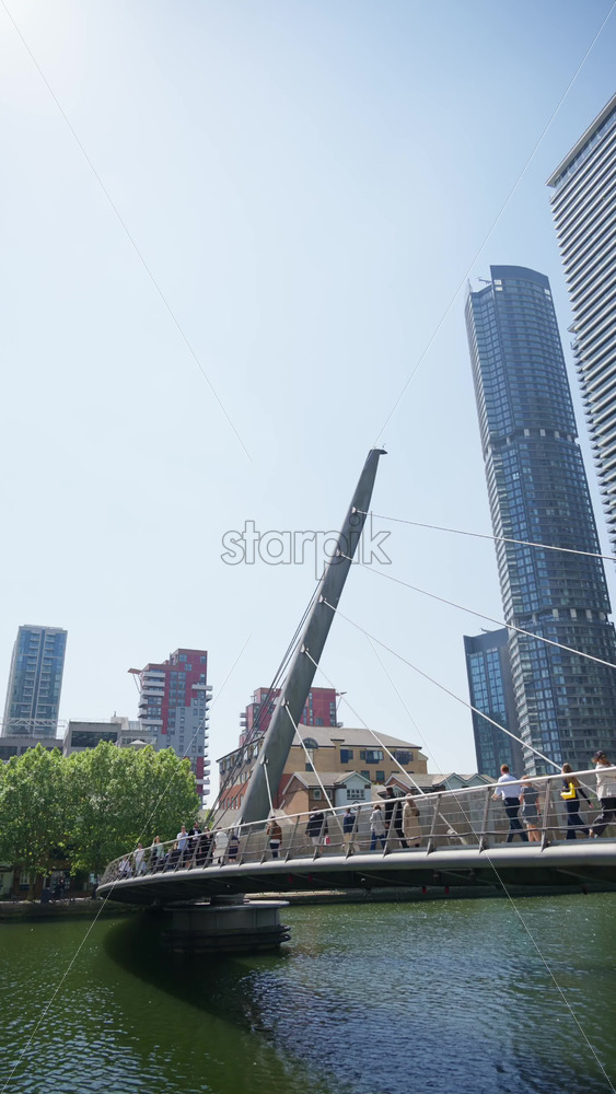 London, England – May 13, 2025: Canary Wharf skyscrapers with people walking across a sleek pedestrian bridge. Vertical - Starpik Stock