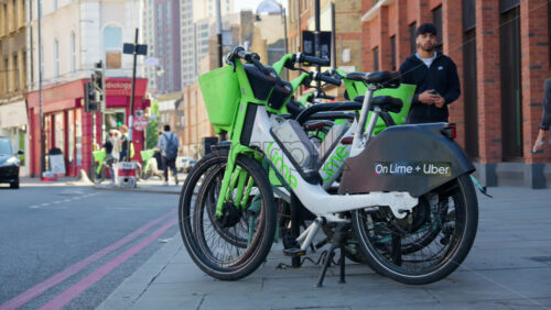 London, England – May 13, 2025: Close-up of Lime and Uber rental bikes parked on street with a cyclist riding by in the background - Starpik Stock