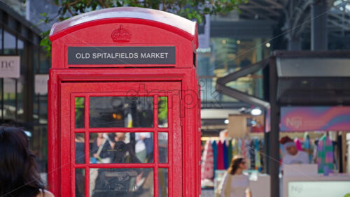 London, England – May 13, 2025: Iconic red British phone booth repurposed with an Old Spitalfields Market sign, set against modern market surroundings - Starpik Stock