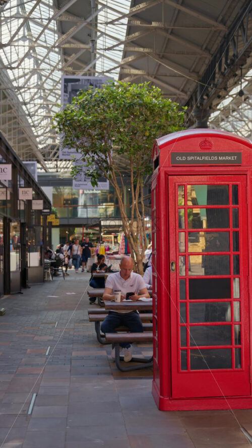 London, England – May 13, 2025: Iconic red British phone booth repurposed with an Old Spitalfields Market sign, set against modern market surroundings. Vertical - Starpik Stock