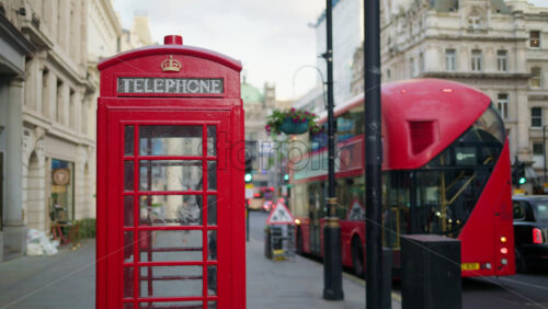 London, England – May 14, 2025: Classic red telephone booth on the street with a moving red double-decker bus in daylight - Starpik Stock