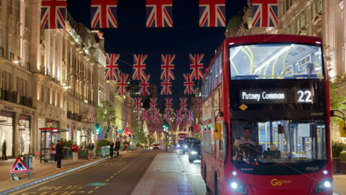 London, England – May 14, 2025: Iconic red double-decker bus driving through Regent street decorated with multiple Union Jack flags at night - Starpik Stock