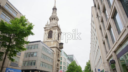 London, England – May 14, 2025: Low angle view of a historic church tower clock nestled between modern and classic buildings on a city street lined with trees and shops in London, England - Starpik Stock