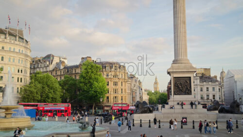 London, England – May 14, 2025: People enjoying a sunny afternoon in the Trafalgar Square with a water fountain, column, and moving red buses - Starpik Stock