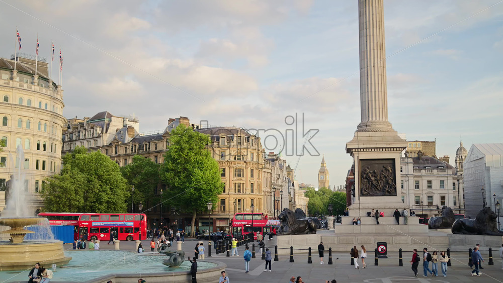 London, England – May 14, 2025: People enjoying a sunny afternoon in the Trafalgar Square with a water fountain, column, and moving red buses - Starpik Stock