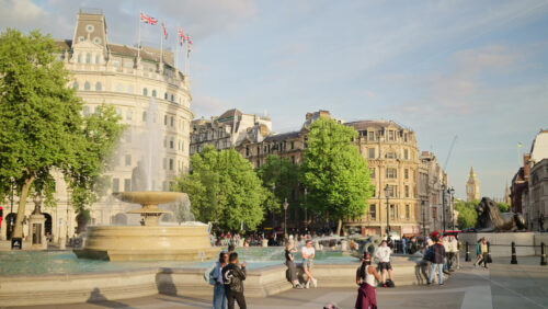 London, England – May 14, 2025: People enjoying a sunny afternoon in the Trafalgar Square with a water fountain, column, and moving red buses - Starpik Stock